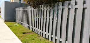 %Title% | Discount Fence Usa A gray wooden fence runs alongside a sidewalk and grassy area with some trees and buildings partially visible in the background.