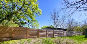 %Title% | Discount Fence Usa Wooden fence with vertical posts runs along a grassy yard, bordered by trees with green foliage under a clear blue sky.