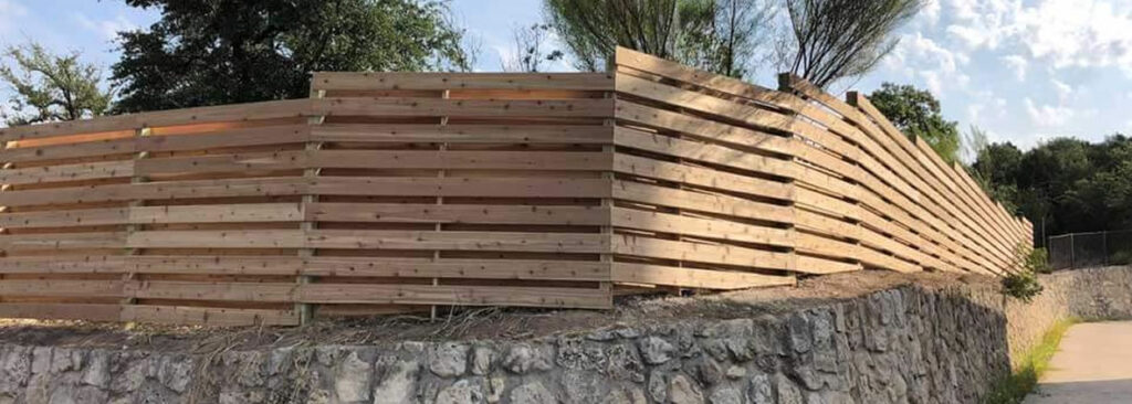 A wooden slat fence stands on top of a stone retaining wall, with trees and blue sky visible in the background.