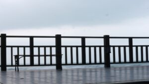 A wooden bench sits on a wet deck beside a railing, overlooking a cloudy, overcast seascape.
