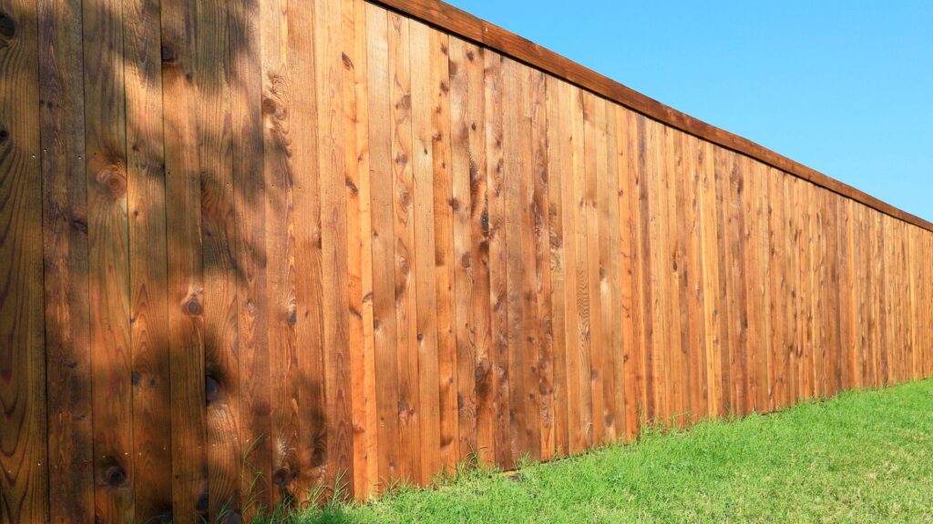 A long wooden privacy fence stands on a green lawn under a clear blue sky, with sunlight casting shadows on the fence.