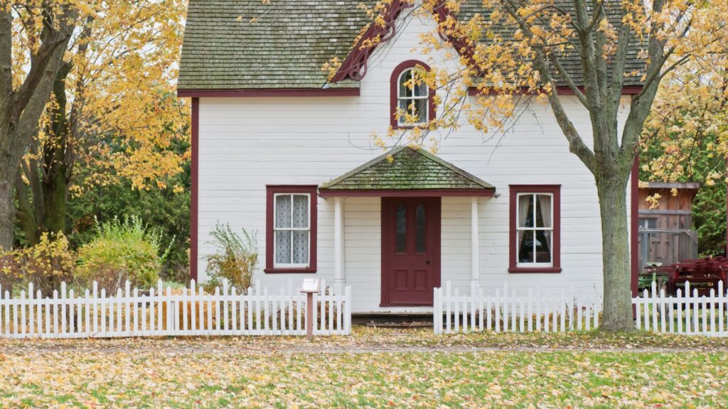 A white house with burgundy trim and a front porch, surrounded by a white picket fence and autumn trees, with fallen leaves on the lawn.