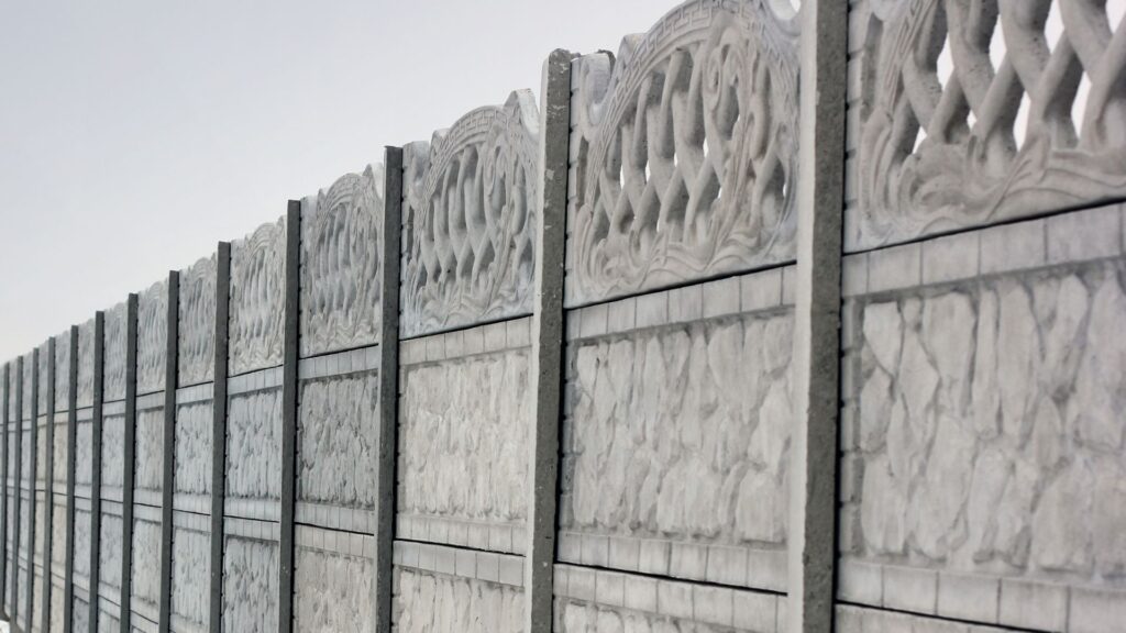 A tall, decorative concrete fence with ornate patterns on the upper panels stands against a pale sky.