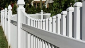 A close-up view of a white vinyl picket fence with pointed tops, lined up in a row outdoors on a sunny day.