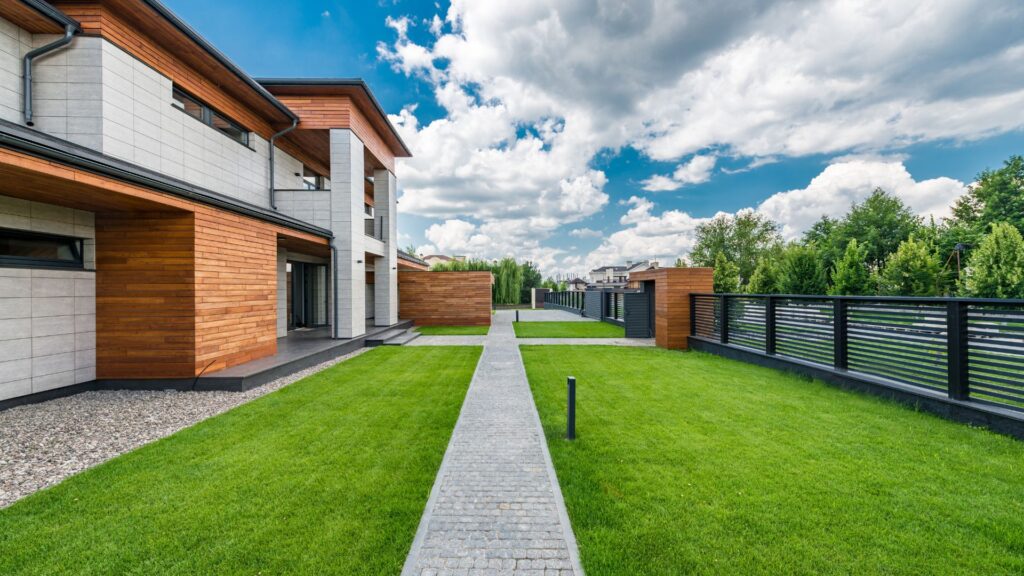 Modern house with wood and concrete exterior, manicured lawn, stone walkway, and metal fence under a partly cloudy sky.