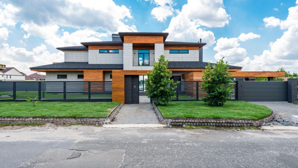 A modern two-story house with wood and concrete exterior, large windows, a flat roof, fenced front yard, and landscaped lawn under a partly cloudy sky.