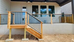 %Focuskw% | Discount Fence Usa A small wooden porch with black metal railings, stairs, and a concrete base is attached to a light-colored house with large windows and a covered roof.