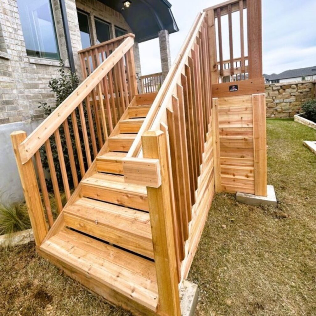 A newly constructed wooden staircase with railings leads from the grass up to the entrance of a brick house.