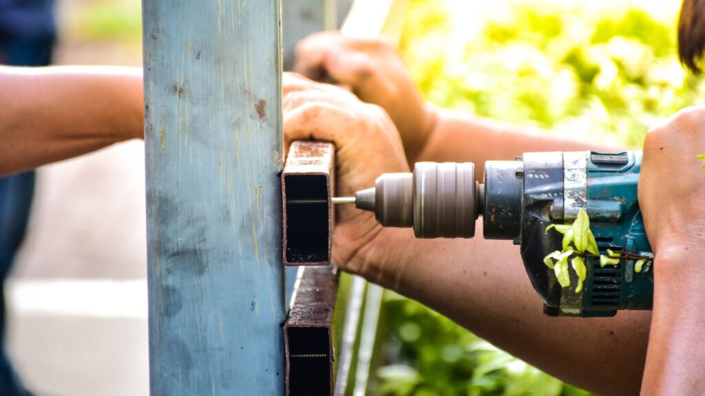 A person drills a hole into a metal square tube while another person holds the tube steady outdoors.
