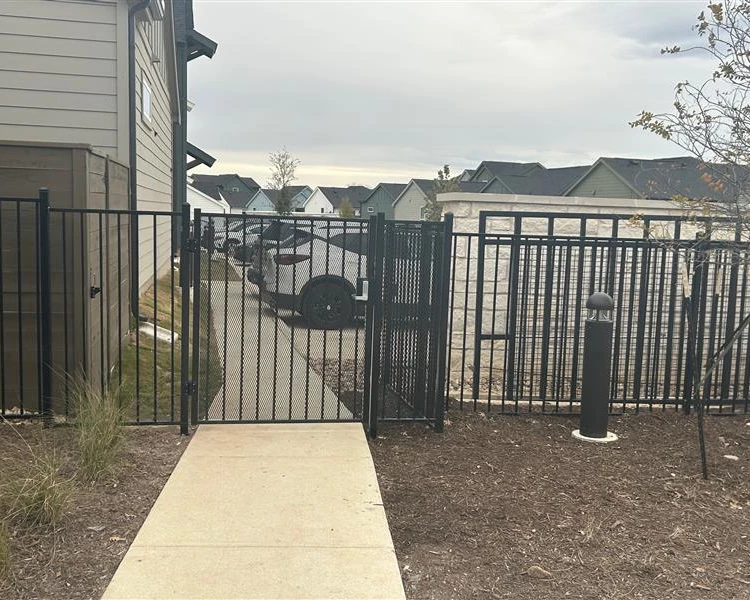 A black commercial metal gate is installed in a fence, but does not align with the adjacent sidewalk, forcing the path around it. Cars and houses are visible in the background.