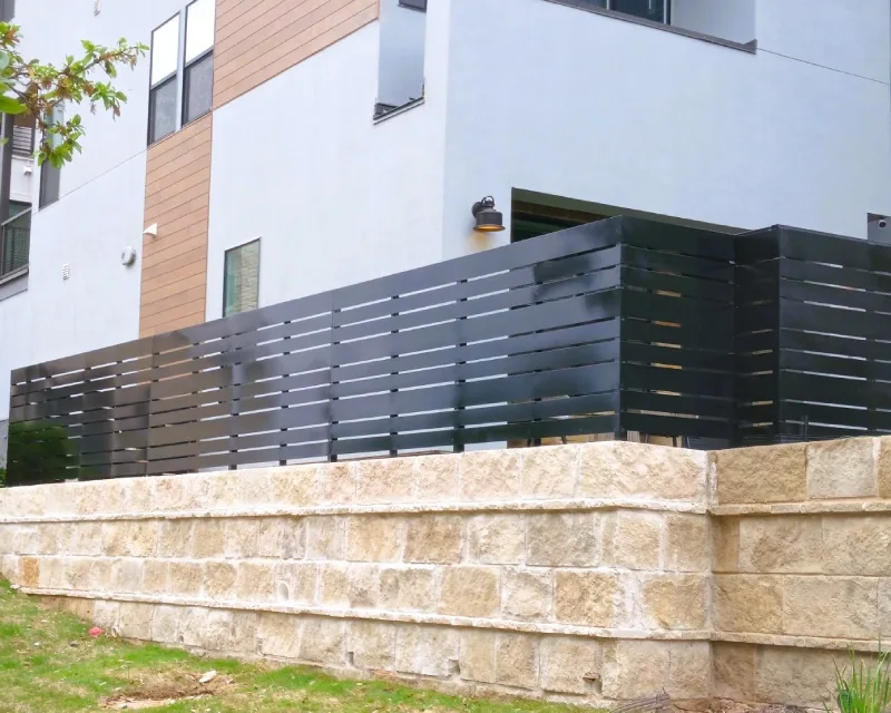 A modern building with a patio enclosed by a sleek privacy fence, featuring horizontal black metal slats atop a light stone retaining wall.
