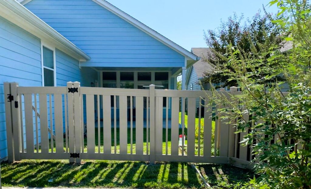 A beige vinyl fence with a gate stands in front of a blue house, surrounded by a grassy yard and some bushes on a sunny day.