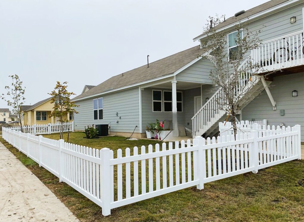 A gray house with a small yard is enclosed by a white vinyl fence. There are a few young trees, and a concrete sidewalk runs alongside the property.