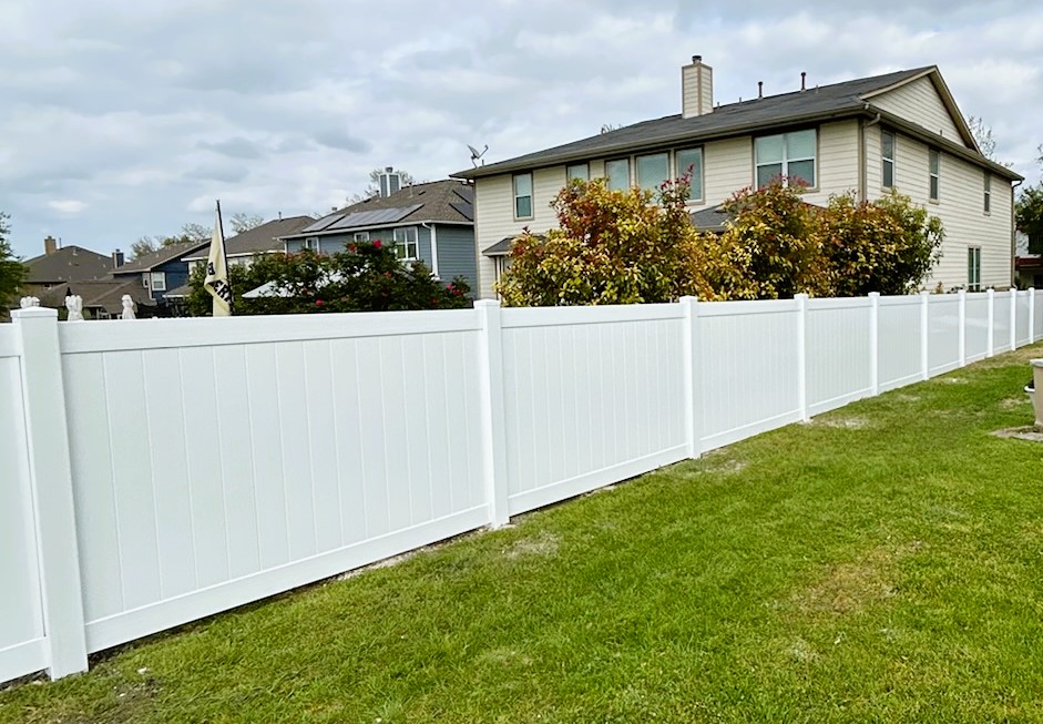A white Vinyl Fence runs along a grassy yard, providing privacy and separating the lawn from neighboring two-story houses surrounded by trees and bushes.