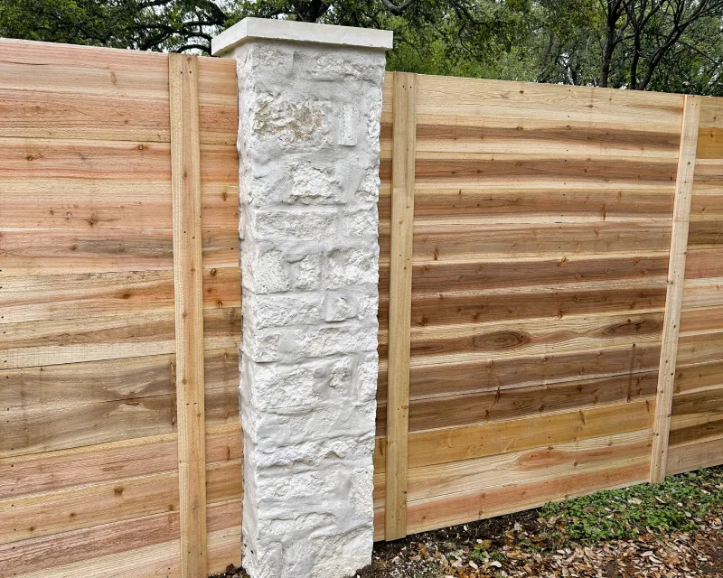 A stone pillar with a white cap stands between sections of wood fencing, with trees and fallen leaves visible in the background.