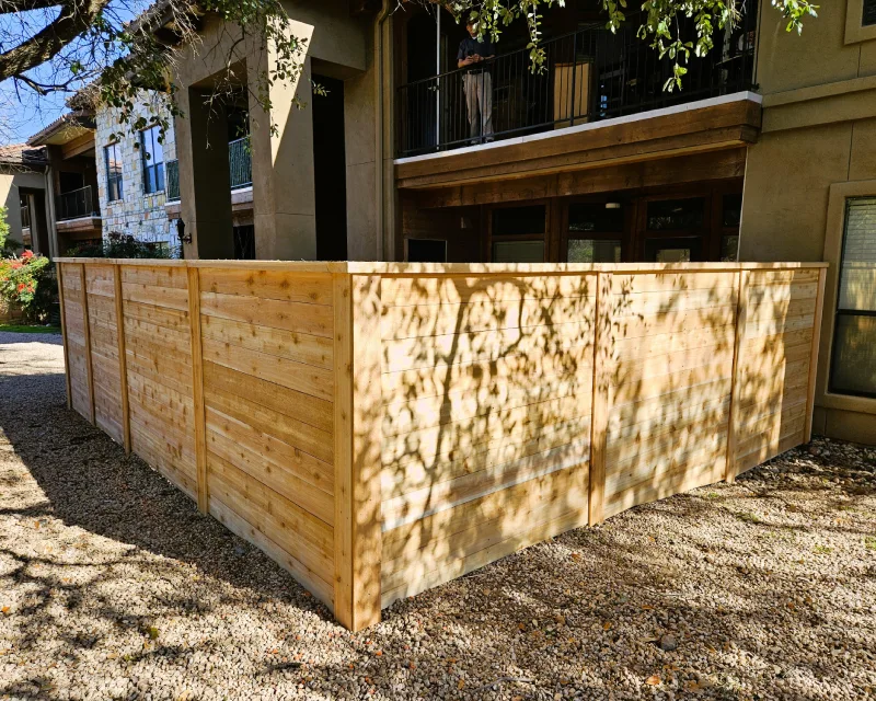 A freshly built wood fencing privacy fence encloses a gravel area next to a residential building, with trees casting shadows on the ground and fence.