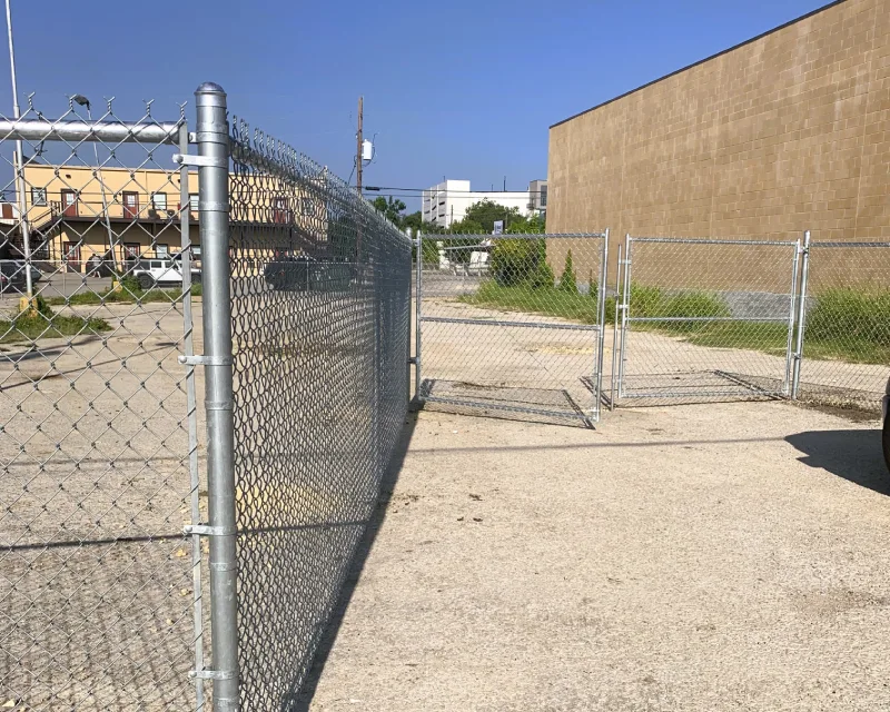 A chain link fence with gates that do not align leaves an open path between the sections in a paved lot beside a building.