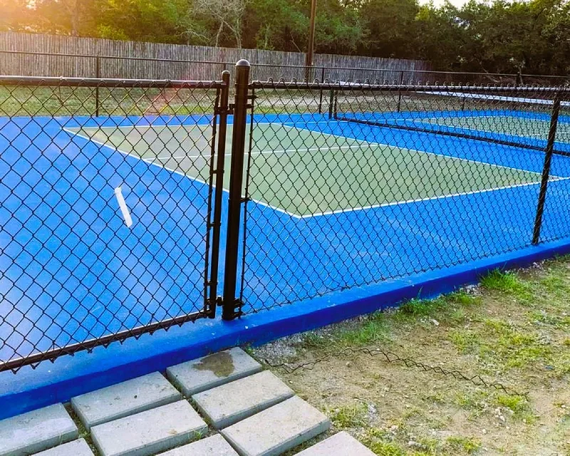 A fenced-in outdoor pickleball court with a blue and green surface, bordered by trees and a chain link fence alongside a wooden fence in the background.
