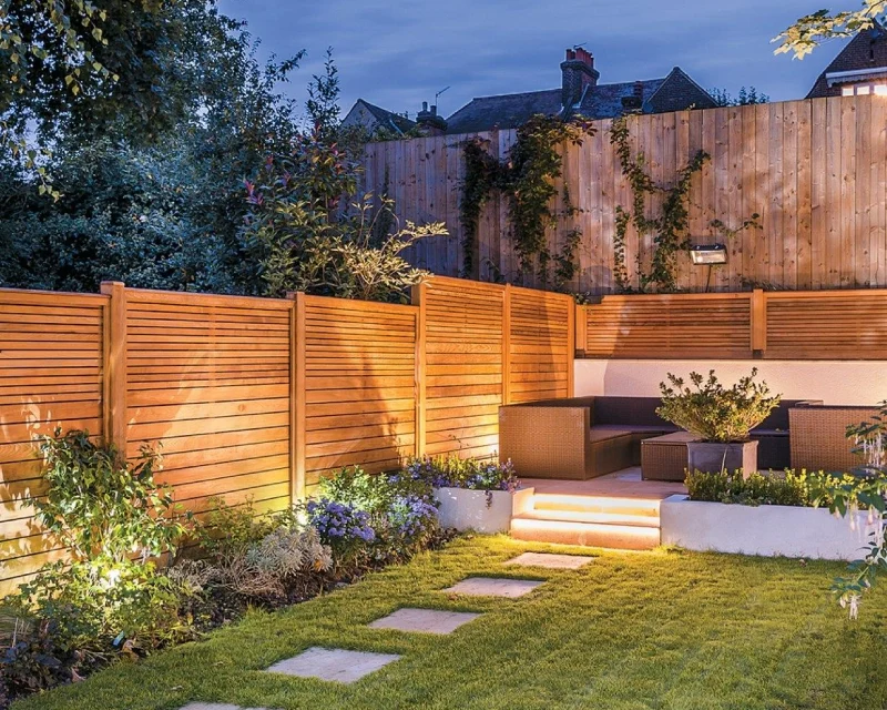 Modern backyard garden with wood fencing, outdoor seating area, lush plants, and soft lighting along steps and fence, leading to a grass lawn with elegant stone pavers.