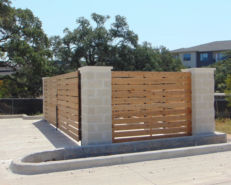 A commercial rectangular enclosure with stone corner pillars and horizontal wooden slats, situated in a paved outdoor area with trees and buildings in the background.