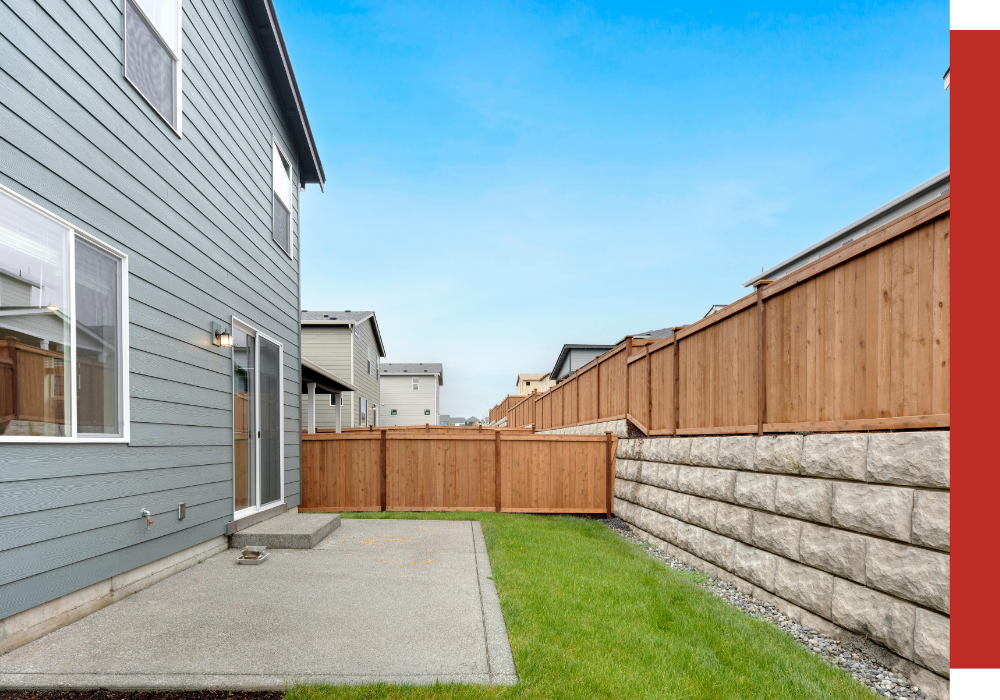 Backyard in Houston with a concrete patio, green grass, wooden fence, and stone retaining wall beside a two-story gray house under a clear sky.