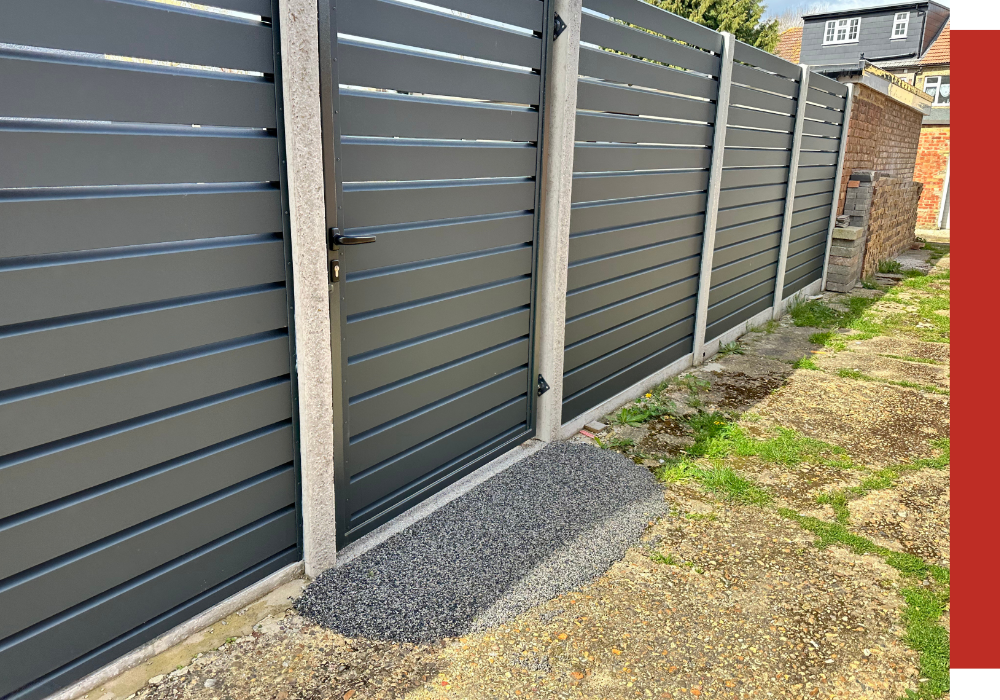 A modern gray metal fence with a gate stands beside a patch of asphalt in Houston, adjacent to a narrow, partly grassy pathway and brick structures.