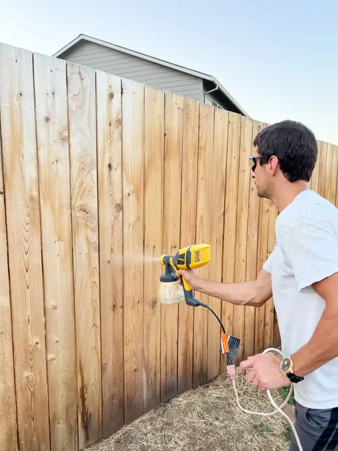A man uses a handheld paint sprayer for fence staining, applying a smooth, even finish to a wooden fence outdoors.