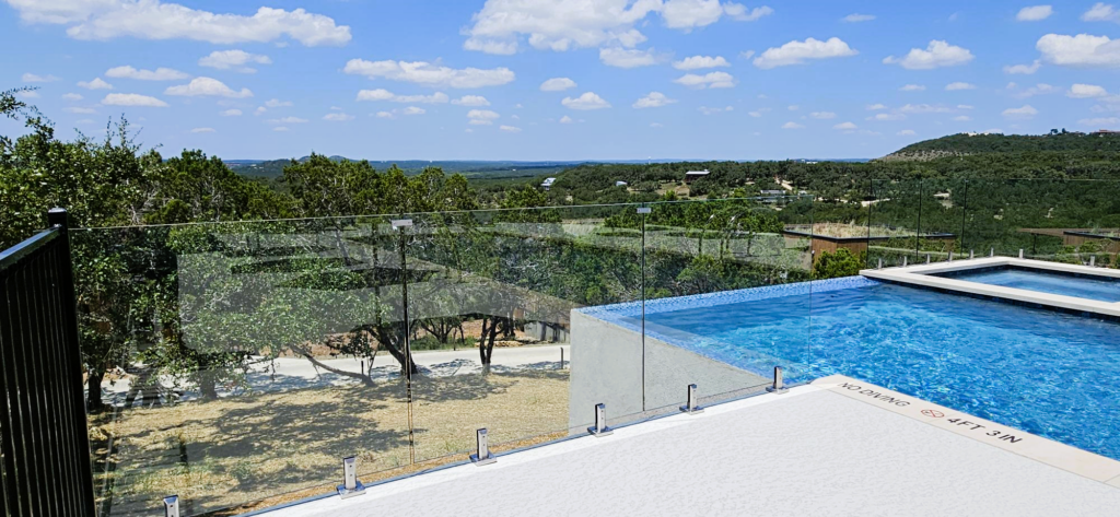 Infinity edge swimming pool with a sleek glass pool fence, overlooking a green, hilly landscape under a partly cloudy sky.