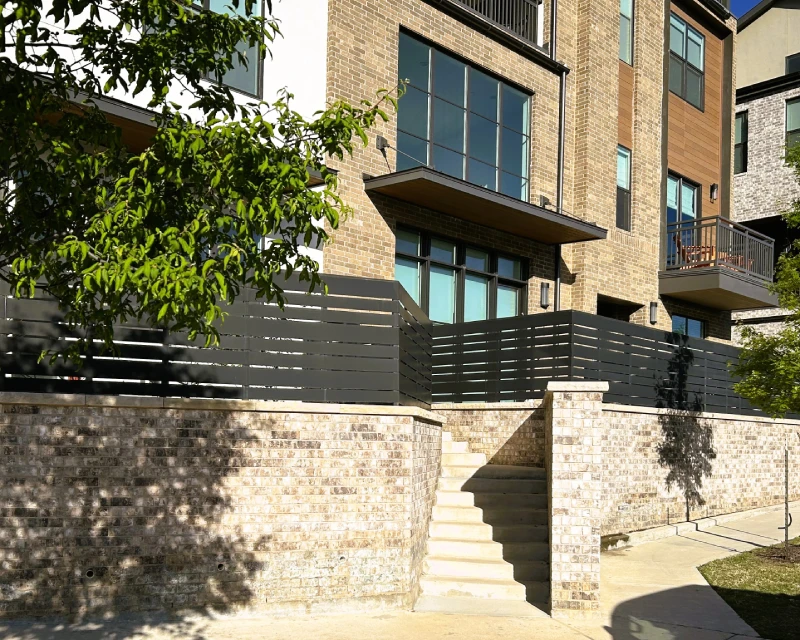 Three-story modern commercial apartment building with large windows, brick exterior, and a staircase leading to a gated patio area enclosed by horizontal metal fencing.