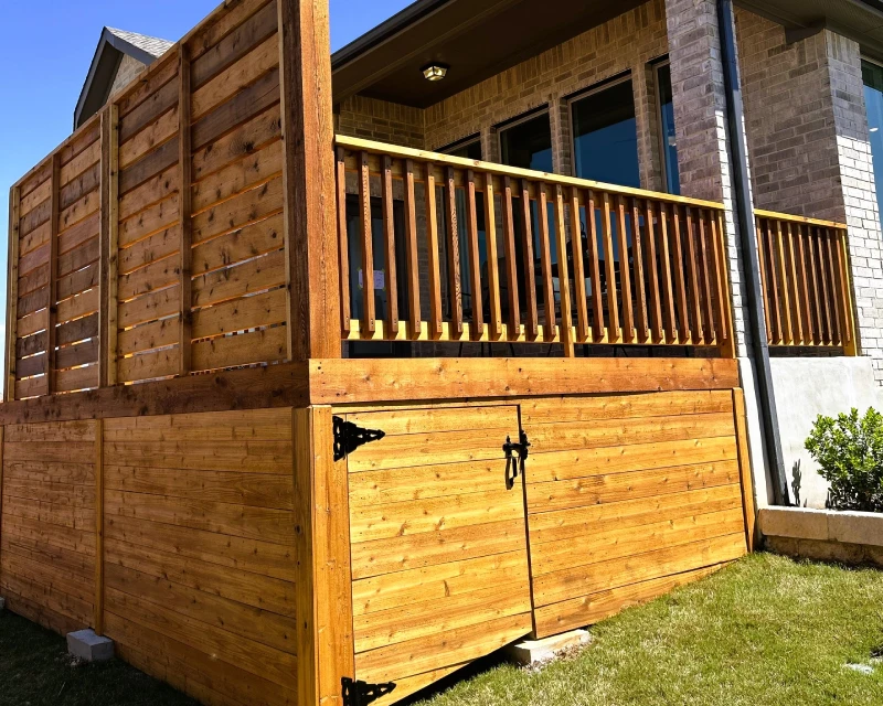 A wooden deck with a privacy fence, railing, and a hinged gate—fresh from deck staining—adjoins a brick house, all glowing in the sunlight.