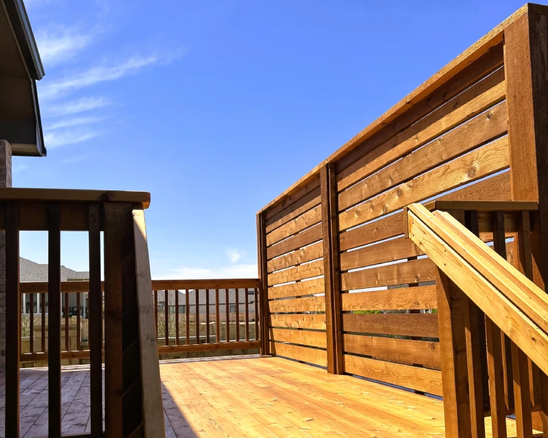 A freshly stained wooden deck with railings and a privacy screen is shown under a clear blue sky, attached to a house—perfect for showcasing expert deck staining.