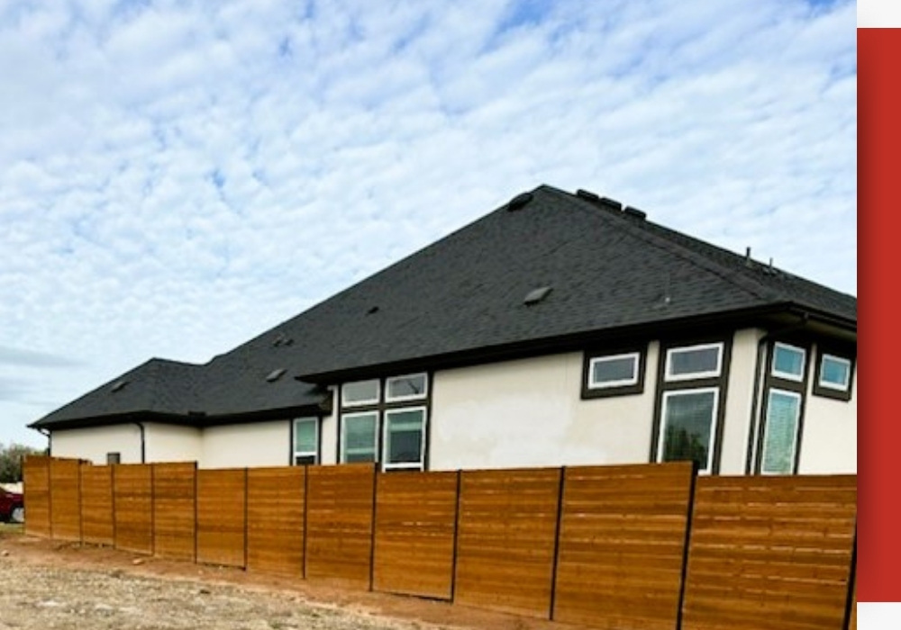 A modern house with a dark roof and large windows in San Antonio is bordered by a tall wooden privacy fence under a partly cloudy sky.