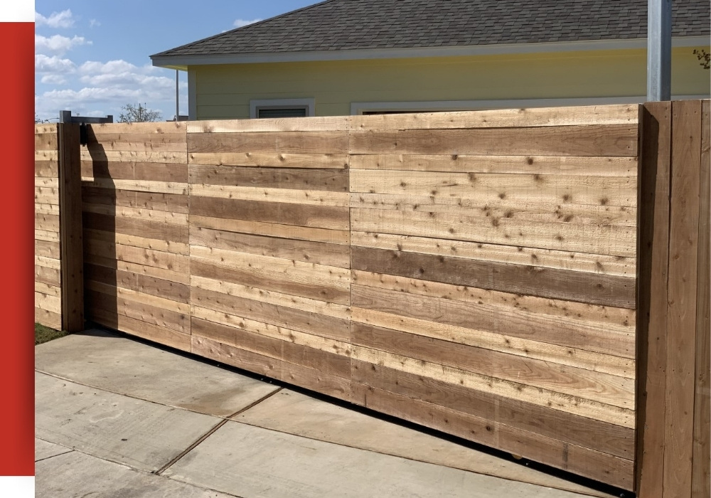 A large, horizontal wooden plank gate stands beside a yellow building with white trim in San Antonio, set on a concrete driveway under a partly cloudy sky.