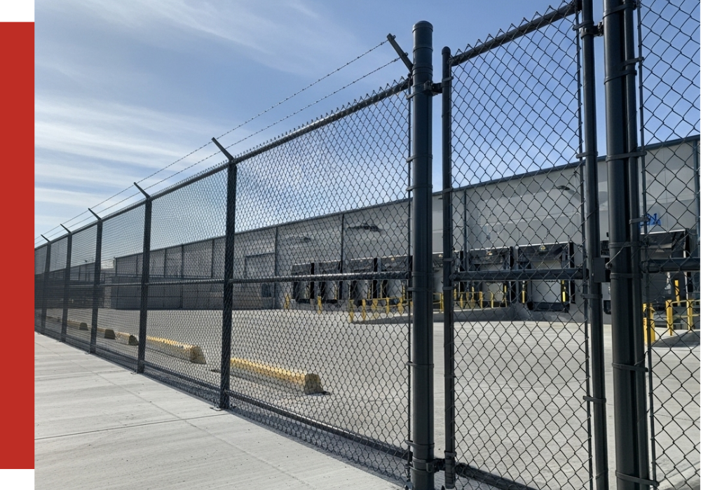 A tall chain-link fence with barbed wire surrounds an industrial loading dock area next to a large warehouse building under a clear blue spring sky.