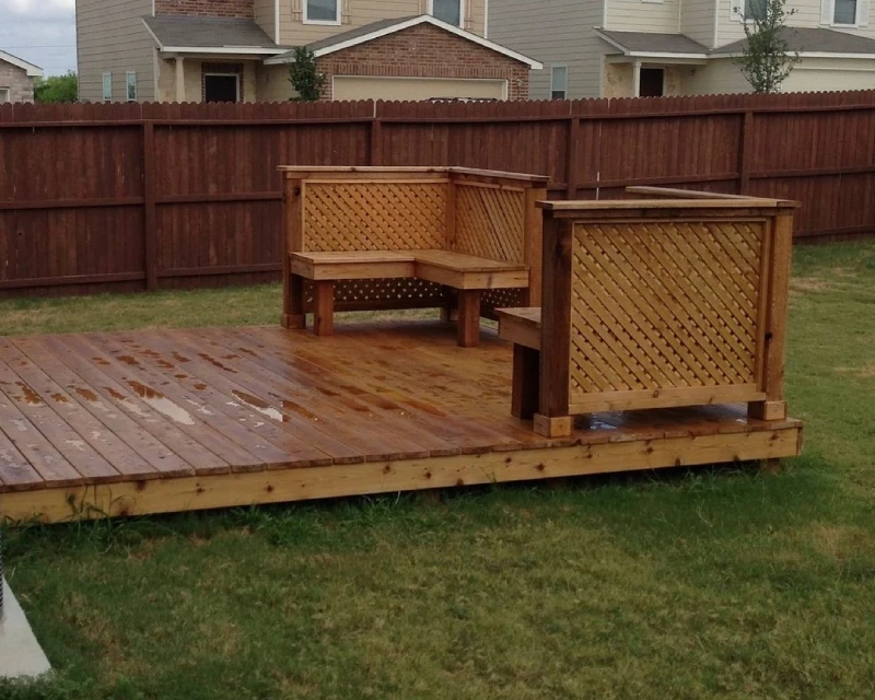 A wooden deck in a backyard features a built-in L-shaped bench with lattice backing, surrounded by grass and bordered by a wooden fence—perfect for relaxing and showcasing your recent deck staining.