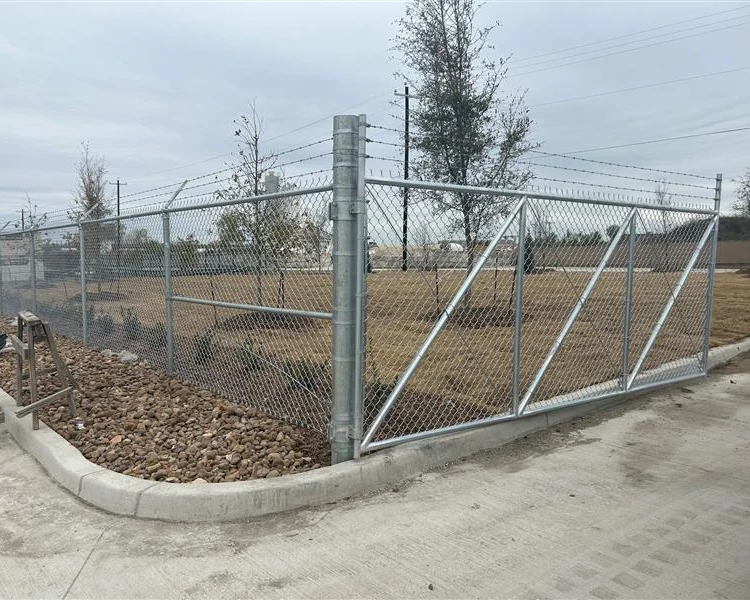 A commercial chain-link fence with barbed wire on top encloses a grassy area with young trees; the gate is installed over a curb, effectively restricting vehicle access.