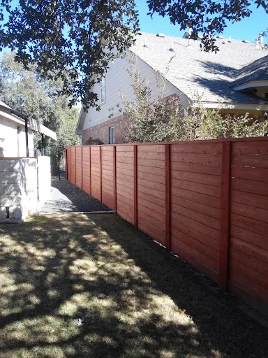 A wooden privacy fence separates two residential houses in South Austin, casting shadows on the grass and gravel below on a sunny day.