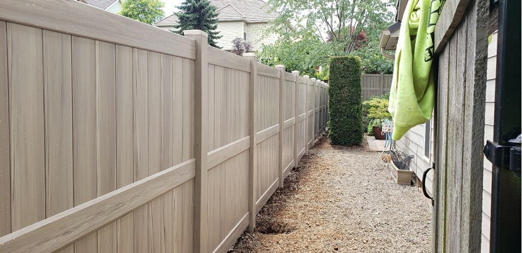 A gravel pathway runs alongside a beige vinyl fence and a house, with plants and a green towel hanging on the right.