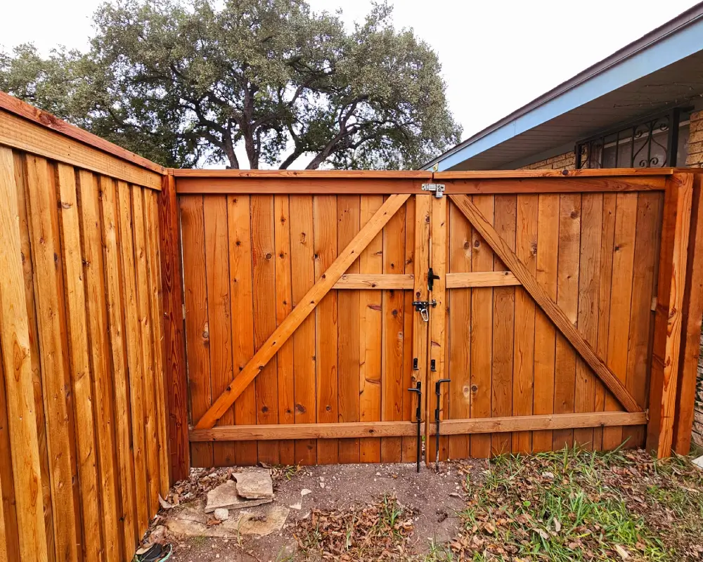 A wooden double gate with metal latches is set in a matching wooden fence, both perfect for showcasing quality fence staining; a tree and part of a house roof are visible in the background.