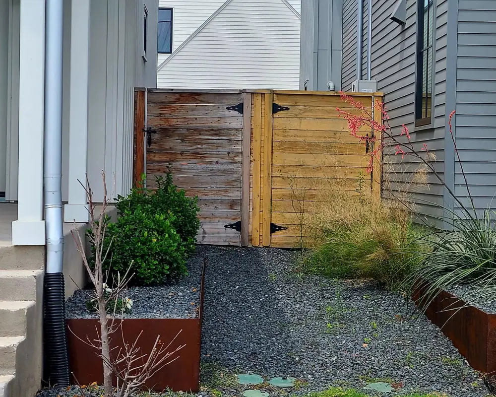 A gravel path leads to a wooden gate between two houses, bordered by metal planters with shrubs, grasses, and a small bare tree—an ideal setting for showcasing expert fence staining.