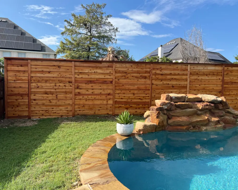 A backyard with a wooden privacy fence featuring fresh fence staining, a small rock waterfall next to the pool, and a potted plant on the grass.