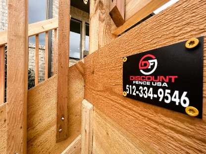 Close-up of a wooden fence with a black and red "Discount Fence USA" sign displaying the phone number 512-334-9546, also offering deck repair services.