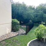 A black metal fence runs alongside a light-colored house, bordering a grassy yard with trees and landscaping rocks.