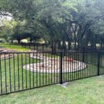 Black metal fence enclosing a grassy yard with circular stone landscaping, trees, and a stone structure in the background.