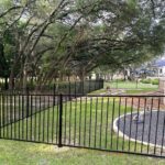 A black metal fence encloses a grassy yard area with trees and a circular gravel section, adjacent to a residential neighborhood.