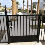 Black metal gate with keypad lock encloses a community pool area, with lounge chairs, palm trees, and houses visible in the background.