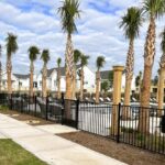 %Imagetitle% | Discount Fence Usa A fenced outdoor swimming pool area with lounge chairs, surrounded by palm trees and residential buildings under a partly cloudy sky.