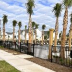A row of palm trees lines a sidewalk next to a black metal fence, with a residential complex and swimming pool area visible in the background under a partly cloudy sky.