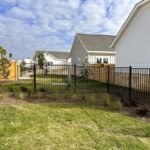 A backyard area with green grass, landscaped plants, and a black metal fence, bordered by light-colored houses and a wooden privacy fence.