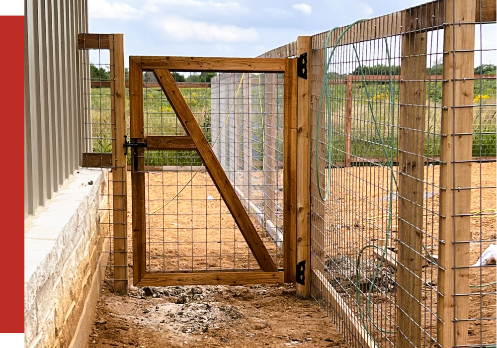 A wooden and wire mesh gate is attached to a wire fence, partially open, with a field visible in the background.
